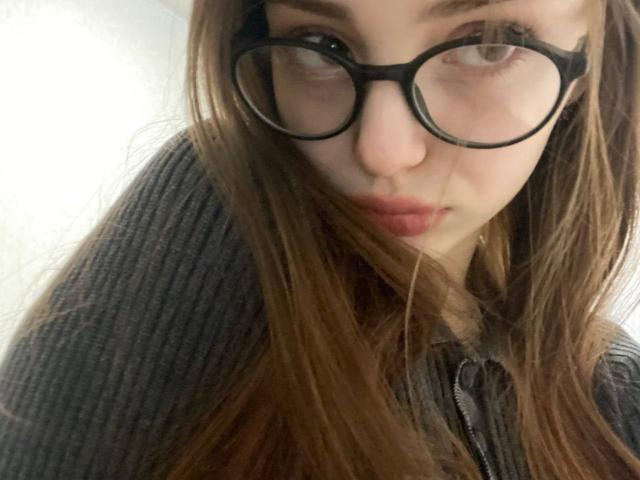 Close-up van Treva Giarro, een jonge vrouw met lang bruin haar, grote zwarte monturen en een serieuze uitdrukking. Ze draagt een gestreept grijze trui. Haar lippen zijn lichtjes open, wat een subtiele seksuele spanning uitstraalt..