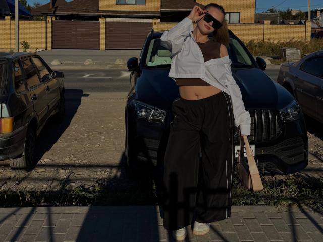 Maragret Gussman è appoggiata a una macchina nera in un parcheggio, indossando una maglietta bianca con una spalla scoperta, pantaloni neri e bianchi. Scarpe da ginnastica. Si sta aggiustando gli occhiali da sole, creando un'ombra sulla sua pancia..