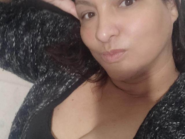 Foto ravvicinata di NinaMilf69, sdraiata, che indossa un maglione nero con una trama particolare e un profondo scollo, che mette in risalto il décolleté. parte di un asfalto nero. Lei sta guardando la fotocamera con un'espressione neutra.