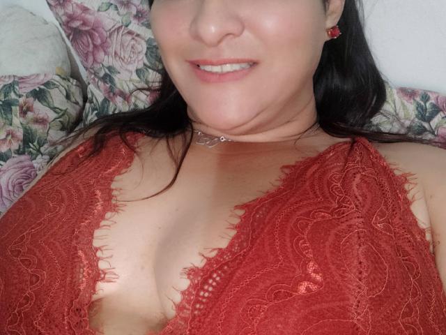 Fotografía de primer plano de NinaMilf69, vistiendo un sujetador de encaje rojo, mostrando el escote, sonriendo ligeramente, cabello negro, piel clara. Almohada con motivos florales en el fondo, sutil tensión sexual, ambiente casual e íntimo..