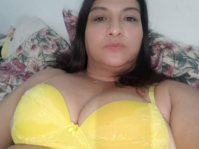 NinaMilf69, une femme corpulente avec de longs cheveux noirs, est allongée sur un lit, portant un soutien-gorge en dentelle jaune vif, et montrant de grandes seins. Coussin floral en arrière-plan. Expression neutre, légère moue..