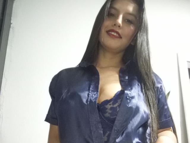 MadissonFontaine, dark-haired woman with red lipstick, wearing a shiny blue satin shirt open to reveal a lace bra. She's smiling slightly, looking directly at the camera, with a strong sexual tension.