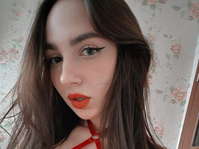 Fotografía de primer plano de Miiraii con piel pálida, cabello marrón oscuro y labios rojos. Ella tiene delineador negro, un collar rojo y una expresión seductora. El fondo es un papel tapiz con motivos florales. Increíblemente sexy..