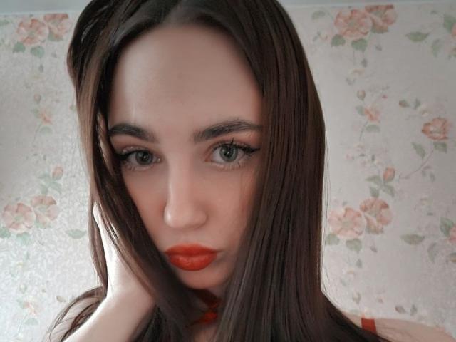 Close-up photo of Miiraii with long, straight brown hair, intense green eyes, and bright red lipstick. Floral wallpaper in background. Miiraii's expression is sultry, with a slight smirk.