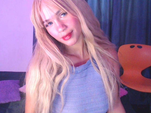 Photograph of LuluDeVine with long blonde hair, blue eyes, and red lips, wearing a light blue top, smiling slightly, with an orange plush toy and dark curtains in the background.