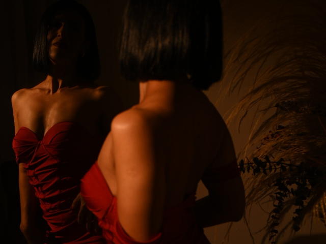 Two women in red strapless dresses, backlit, creating a sultry, shadowy effect. Both have short black hair. One woman's arm is around the other's waist, enhancing the intimate, sexual tension.