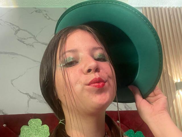 Close-up photo of GraceRedMiller, wearing a green hat, with glittery green eyeshadow, red lips, and dark brown hair. She's playfully pouting, hand on hat, green confetti in the background.