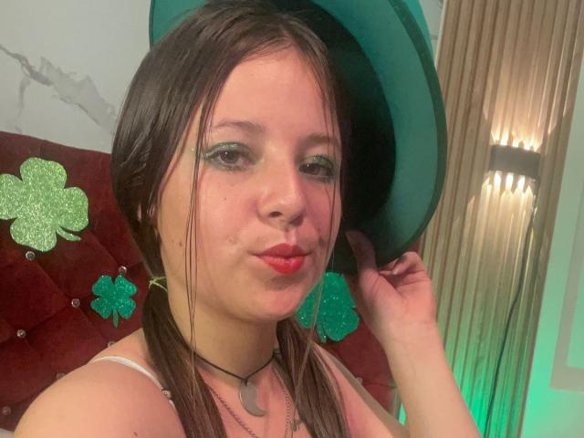 Photographie de GraceRedMiller avec des cheveux bruns lisses, portant un chapeau vert, du rouge à lèvres rouge et un collier noir. Des décorations en forme de trèfle vert sont derrière elle. Elle fait la moue, et sa main touche son chapeau. Éclairage lumineux.