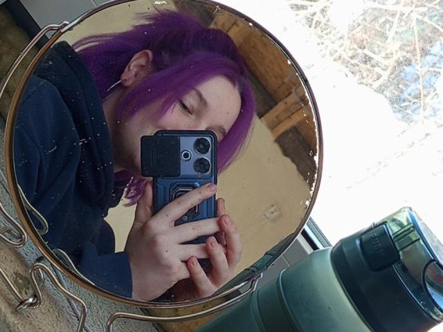 Spiegelfoto van AnnitaMilliter met paars haar, ogen gesloten, die een zwarte telefoon vasthoudt, gekleed in een zwarte top, in een zwak verlichte omgeving. kamer, met een groene waterfles aan de rechterkant, die een serieuze uitdrukking weerspiegelt..