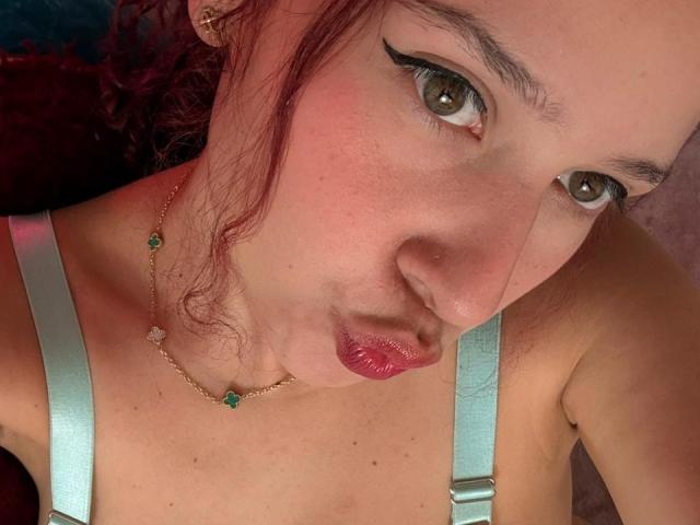 Close-up of ValeryScot, pouting with glossy red lips, brown eyes, and heavy black eyeliner. She wears a silver top with straps and a delicate necklace. Her skin is fair and freckled.