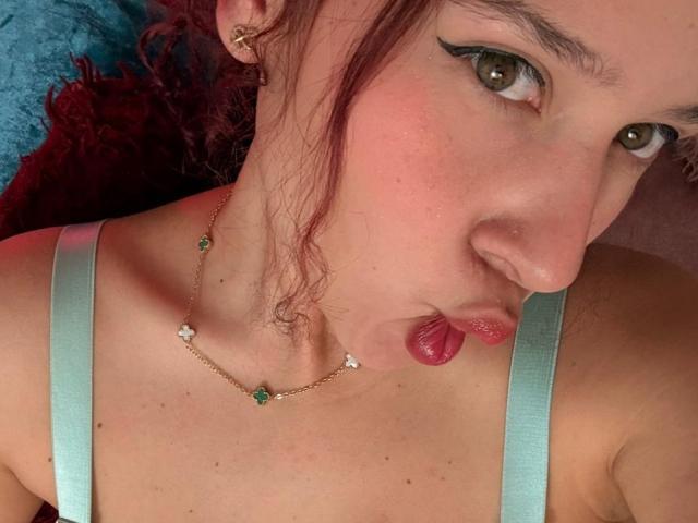 Close-up of ValeryScot, pale-skinned woman with red hair, black eyeliner, and red lipstick, pouting with tongue out. She wears a light green strap top and a gold necklace with small jewels.