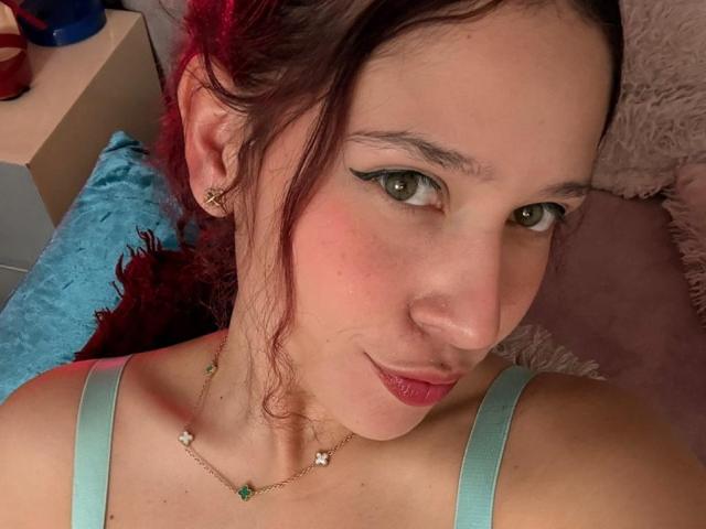 Close-up of ValeryScot, a young woman with green eyes, red lipstick, and dark brown hair, wearing a light green tank top and a delicate gold necklace. Sexual tension, subtle smirk.