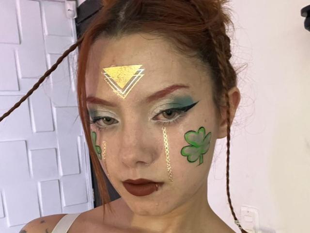 Primo piano di Emma Thompson con il trucco: trifogli verdi sulle guance, un triangolo giallo sulla fronte, sopracciglia rosse, occhi blu-verdi. ombretto, rossetto scuro.
È rivolta verso il basso, con uno sfondo di un muro di mattoni bianchi. Incredibilmente sexy..