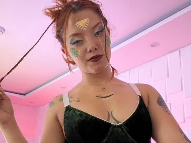 Fotografía de Emma Thomson con cabello rojo recogido en un moño despeinado, vistiendo una blusa negra, con maquillaje en la cara que incluye círculos verdes y un triángulo. frente, sosteniendo un palo, tatuajes en los hombros, tensión sexual.