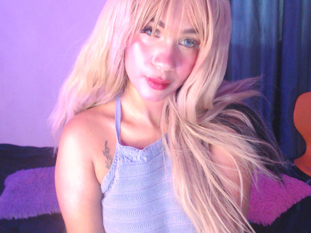 Fotografía digital de LuluDeVine con cabello rubio platino, ojos azules y una camiseta blanca. La iluminación brillante resalta su rostro, con un fondo suave en tonos rosa y negro. Increíblemente atractivo/a..