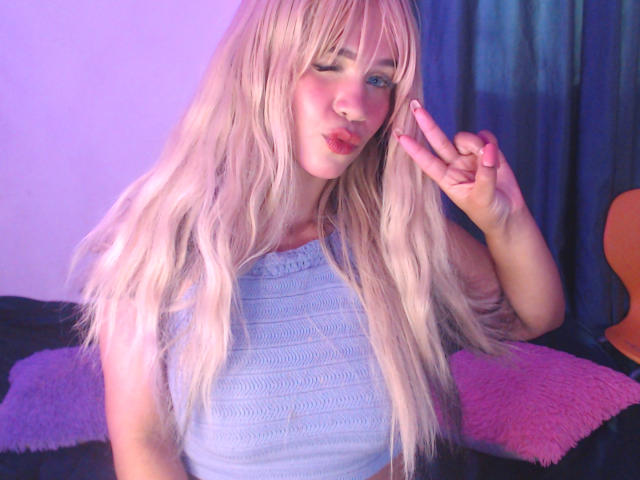 LuluDeVine, blonde, wavy hair, blue eyes, wearing white textured top, winking, pink lips, hand up, dark blue and pink background, bright lighting, sexual tension, casual pose, vibrant colors.