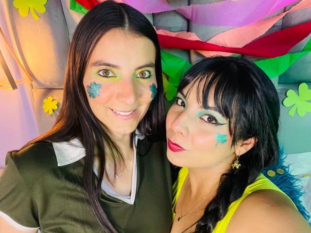 ValkiriaAndDione, deux femmes aux cheveux noirs, souriant à l'appareil photo. L'une porte un haut vert, l'autre un haut jaune, et toutes les deux ont du maquillage bleu en forme d'étoile sur le visage. Fond clair et coloré. Extrêmement proche..