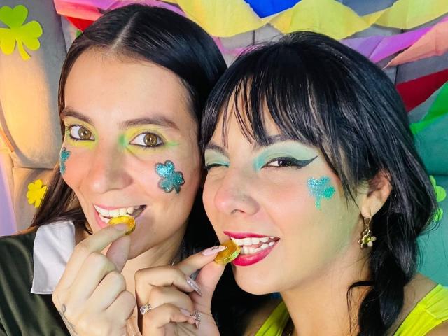 Fotografia ravvicinata di ValkiriaAndDione, due donne con capelli neri, trucco colorato sul viso e motivi floreali, che tengono caramelle. nelle loro bocche, sorridenti, colori vivaci, giocosi, tensione sessuale, sfondo colorato.