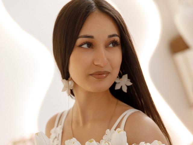 Fotografia de Elvira Wesdety com cabelos longos e escuros, vestindo um vestido branco com estampas florais e brincos de flores combinando, olhando para o lado com um sorriso discreto, fundo branco claro. Absolutamente deslumbrante..