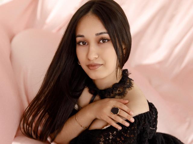 Fotografie von Elvira Wesdety, einer jungen Frau mit langen, schwarzen Haaren, die einen schwarzen Spitzen-Choker und ein schulterfreies Kleid trägt. Sie sitzt auf einem rosa Sofa, ihre Hände liegen gefaltet vor ihrer Brust, und sie blickt direkt in die Kamera, mit einem leichten Lächeln. Sie trägt einen schwarzen Ring an ihrer rechten Hand. Sanftes Licht betont ihre glatte, helle Haut. Sexuelle Spannung ist vorhanden, was durch ihren direkten Blick und ihre leicht geöffneten Lippen betont wird..