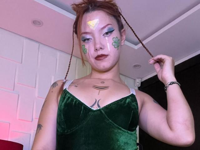 Photographie d'Emma Thomson avec des cheveux tressés, une robe en velours vert, et du maquillage sur le visage avec des trèfles verts et un triangle jaune. Tatouages visibles sur la poitrine, cheveux tressés, tension sexuelle, pensées à propos du pénis..