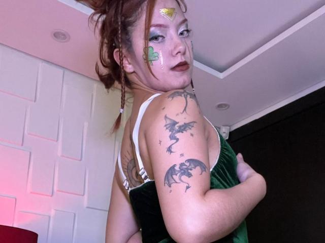 Emma Thompson, con un tatuaje de dragón y estrellas en el brazo, tirantes de sujetador blancos, vestido verde oscuro, rostro pintado con dorado y verde. y manchas rojas, mirando por encima del hombro, sexy, ligeramente girada, en un ambiente interior..