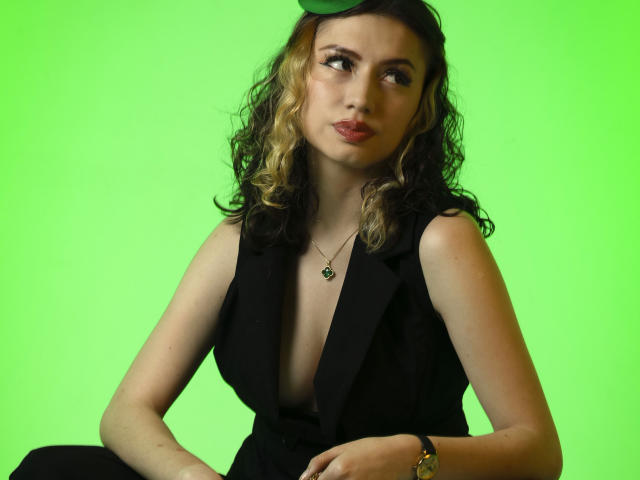 Photograph of ElizaCanterville with curly brown hair, wearing a black sleeveless dress with a deep neckline, green background, green hat, gold bracelet, and necklace, looking to the side, sexy.
