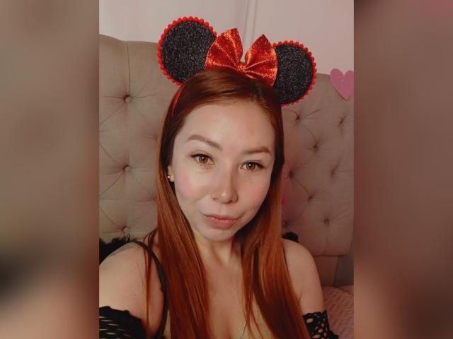 Fotografie von MiraDulce, einer asiatischen Frau mit langen roten Haaren, die schwarze Minnie-Maus-Ohren mit Glitzer und eine rote Schleife trägt, schwarz top mit offener Schulter, gesteppte, beige Kopfteil, rosa Herz-Dekoration. Verdammt süß, sexy..