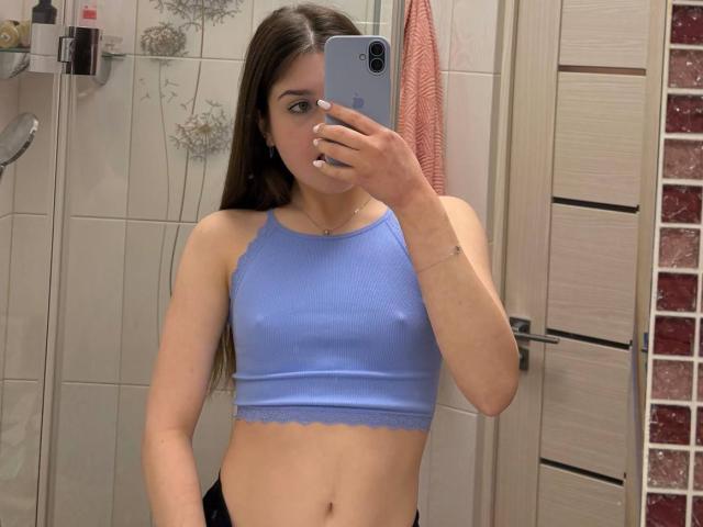 CristenStamper with long dark hair, wearing a tight blue sports bra, holding a phone in front of her face, mirror selfie. Bathroom background with towels and tiles. Focused on her toned midriff and casual pose.