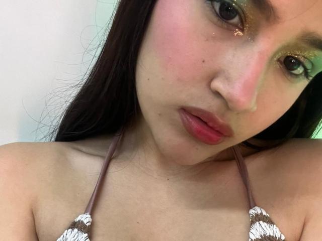 Close-up of IrisBlair, Asian woman with long black hair, green glitter eye shadow, pink lips, and red lipstick. She's looking directly at the camera with a slightly open mouth. Topless, halter straps visible.