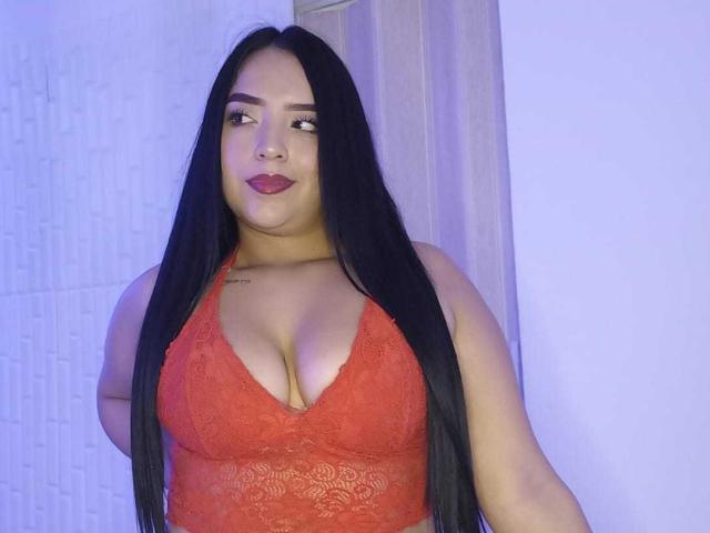 AdaraCute69, com cabelos longos e pretos, vestindo um top de renda vermelho com alças, mostrando bastante decote, olhando para o lado com batom vermelho escuro, iluminação com tons de azul, leve tensão sexual.