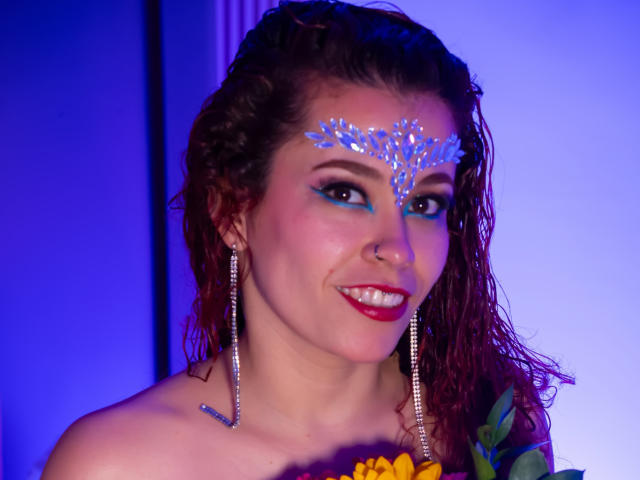 Nahaufnahme von Arielle Brooks mit nassen, braunen Haaren, die glitzernde blaue Gesichtsbemalung auf der Stirn trägt, mit silbernen Ohrringen und roten... Lippenstift und ein trägerloses Oberteil, mit einem verführerischen Lächeln, blaues und violettes Licht..