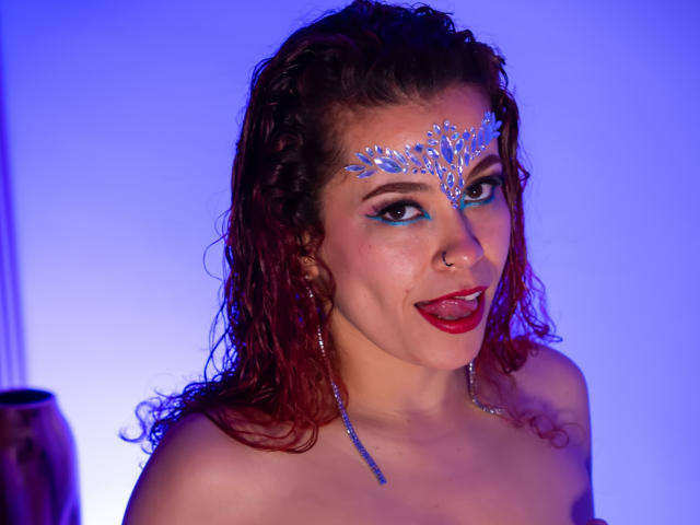 Fotografia di Arielle Brooks con i capelli castani ricci e bagnati, che indossa un trucco per il viso blu e un copricapo argentato. con la lingua fuori, capezzolo visibile, sfondo blu, espressione seducente..