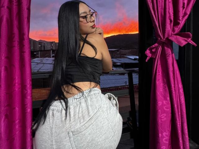 ChloeMoretti stands with her back to the camera, long black hair, wearing glasses, black crop top, and white loose pants. Sunset sky with orange and pink hues, purple curtains. Sexual tension.