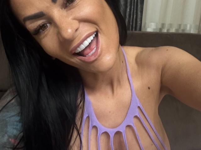 Close-up of AndraD with dark hair, smiling with tongue out, wearing a purple halter top with cutouts revealing cleavage. Shiny skin, brown eyes, and visible moles on shoulders. Fucking sexy.