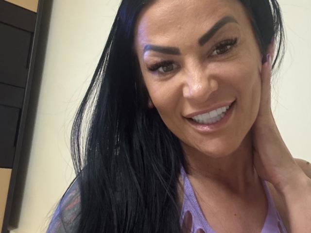 Photo en gros plan d'AndraD avec de longs cheveux noirs, souriant avec des lèvres brillantes, des sourcils épais, et une main derrière sa tête. Elle porte un haut violet clair et regarde directement l'appareil photo. Extrêmement sexy.