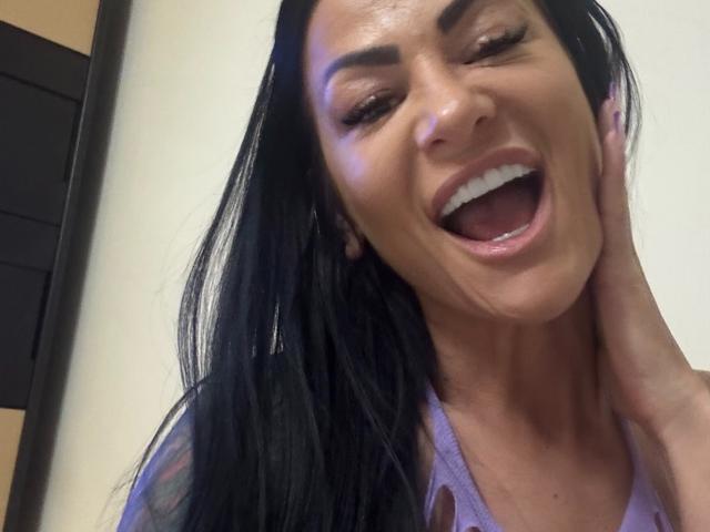 Gros plan d'AndraD, une femme séduisante avec de longs cheveux noirs, souriant largement, la bouche ouverte, une main touchant sa joue. Elle porte un haut lavande, elle a l'air excitée et séduisante..