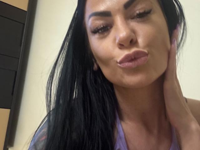 Photo en gros plan d'AndraD, une femme aux longs cheveux noirs, aux lèvres pulpeuses et aux sourcils épais, qui fait légèrement la moue tout en touchant quelque chose. Elle passa sa main sur sa joue. Son expression est sensuelle et séduisante..