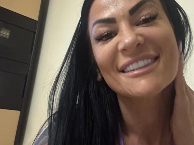 Fotografía de primer plano de AndraD con piel brillante, cabello negro y una sonrisa amplia que muestra sus dientes. La mano de AndraD descansa sobre su cuello, mostrando cejas gruesas y arqueadas y labios carnosos. Tensión sexual evidente..