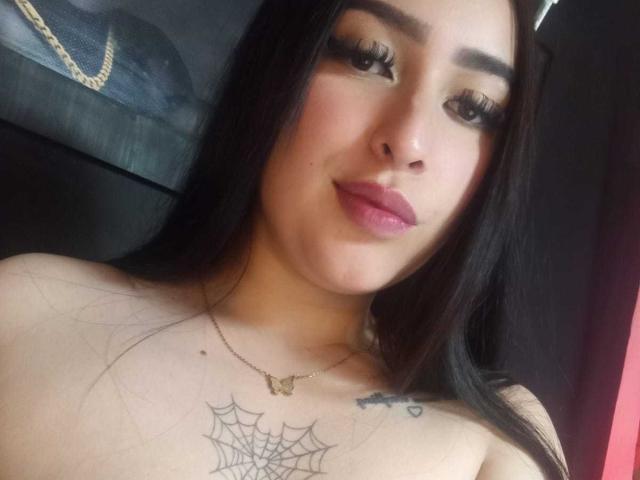 Fotografia ravvicinata di Chloey69, una donna con lunghi capelli neri, un trucco occhi molto marcato e rossetto rosa, che mostra un tatuaggio a forma di ragnatela. sul suo petto, senza top, con il seno scoperto e una collana d'oro..