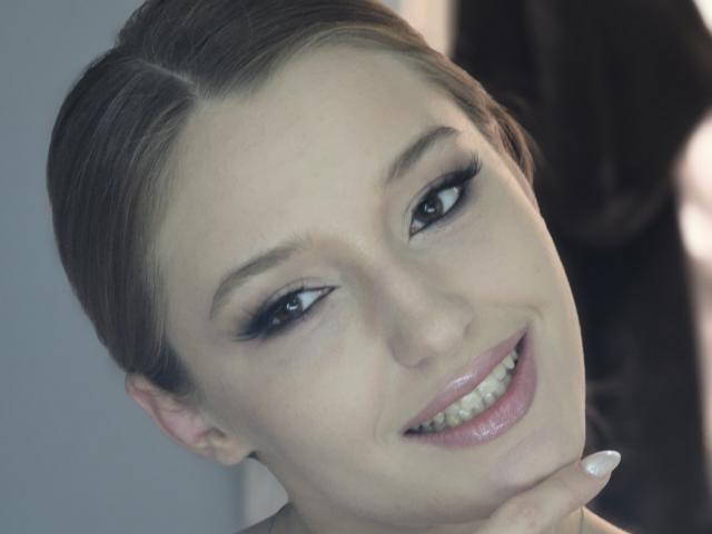 Foto em close-up de DiedraBloschichak, com pele clara, cabelo castanho preso para trás e um sorriso largo mostrando lábios brilhantes. Ela tem uma linha de delineador preto bem marcada, unhas brancas e uma leve inclinação na cabeça. A expressão dela é sedutora e sensual..