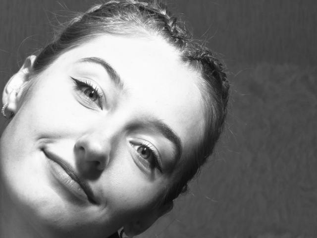 Black and white photo of DiedraBloschichak's face, tilted to the right. Bright highlights on her forehead and cheek contrast with dark shadows. She's smiling subtly, with a hint of sexual tension.