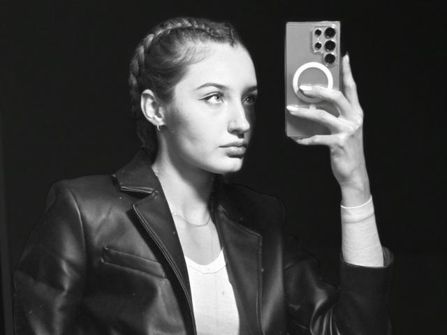 Foto em preto e branco de DiedraBloschichak, uma mulher com cabelo trançado, vestindo uma jaqueta de couro, tirando uma selfie com telefone. Sua expressão é séria, capturando seu perfil. Alto contraste..
