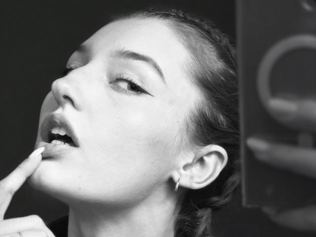 Gros plan en noir et blanc de DiedraBloschichak, les yeux fermés, la tête penchée en arrière, et un doigt dans la bouche. Le contraste élevé et la lumière douce mettent en valeur ses traits et ses cheveux foncés. Tension sexuelle évidente..