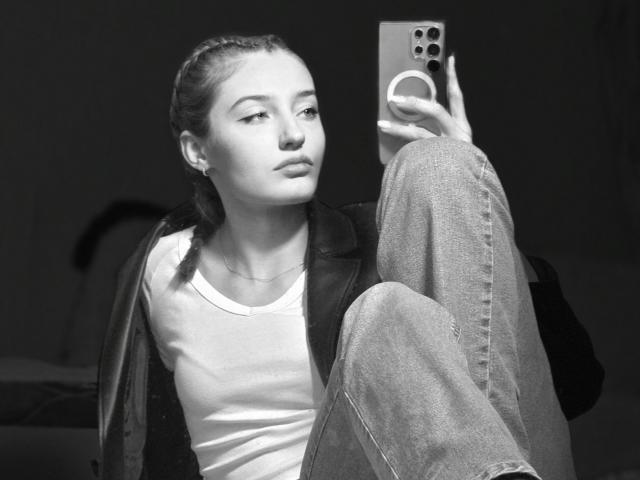 Fotografia em preto e branco de DiedraBloschichak sentada com uma perna levantada, segurando um smartphone na mão direita. Ela veste uma camiseta branca e calças jeans, e está olhando para o celular. Pose incrivelmente sensual..