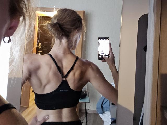 Diedra Bloschichak, met een gespierde rug en duidelijke schouderbladen, draagt een zwarte sportbeha en maakt een selfie. spiegel. Het scherm van haar telefoon straalt fel in haar rechterhand..