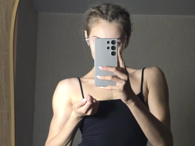 Fotografía de Diedra Bloschichak, sosteniendo un iPhone blanco con tres lentes de cámara, que le ocultan el rostro. Ella lleva una camiseta negra con tirantes finos y se está tomando una foto frente al espejo. Su cabello está trenzado. Una luz brillante resalta sus brazos tonificados..