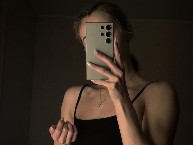 DiedraBloschichak, em um quarto escuro, tira uma selfie com um iPhone, cobrindo o rosto com o aparelho. Ela está vestindo uma blusa preta com alças finas e segurando o telefone com uma mão. As unhas dela são longas e brancas. A iluminação realça seus ombros nus e a clavícula..