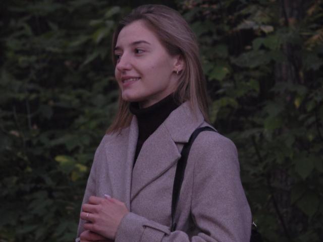 Diedra Bloschichak, avec des cheveux bruns mi-longs, souriant légèrement, portant un manteau gris par-dessus un pull noir à col roulé, les mains serrée sur le devant, sur un fond de feuilles vert foncé, une tension sexuelle subtile..