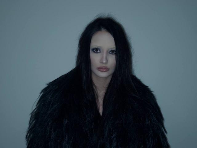 MarchelleBerrien, a pale-skinned woman with striking blue eyes, stares directly at the camera. She's wrapped in a shaggy black fur coat, creating a sexy, mysterious vibe. Blue-tinted lighting enhances the effect.
